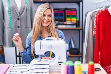 Blonde woman dressmaker designer using sew machine looking away to side with smile on face, natural expression. laughing confident.