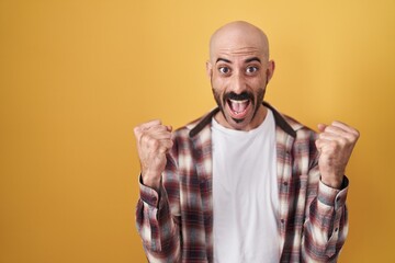 Naklejka premium Hispanic man with beard standing over yellow background celebrating surprised and amazed for success with arms raised and open eyes. winner concept.