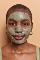Self care and beauty. Serious short haired African woman with dark skin applies nourishing clay mask on face for reducing fine lines wears goldern earrings has well cared body isolated over brown wall