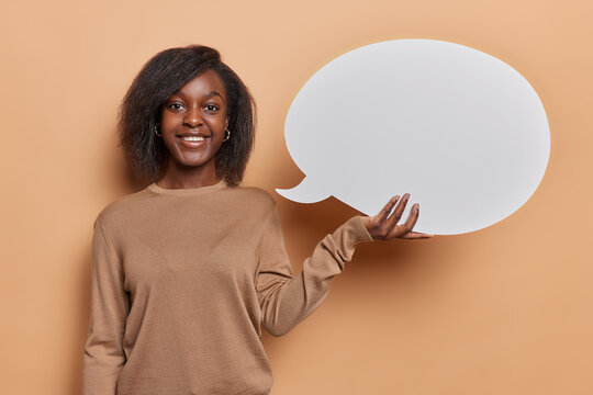 Horizontal shot of young African woman with dark skin gives advice and shares her thoughts dressed in casual jumper smiles broadly isolated over brown background. Empty word cloud. Idea conept