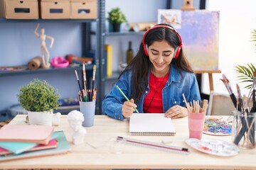 Young hispanic girl artist listening to music drawing on notebook at art studio