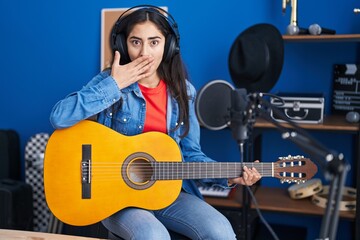 Young teenager girl playing classic guitar at music studio covering mouth with hand, shocked and afraid for mistake. surprised expression