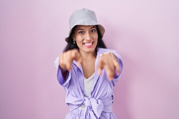 Young hispanic woman standing over pink background wearing hat pointing to you and the camera with fingers, smiling positive and cheerful