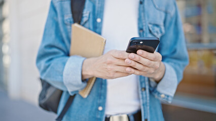 Young hispanic man student using smartphone holding book at university