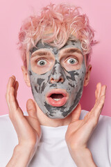 Shocked scared man applies nourishing clay mask on face has dry skin need deep cleansing stares amazed has mouth widely opened clasps hands stares with eyes popped out wears t shirt isolated on pink