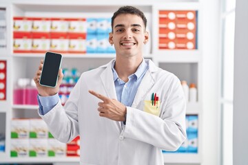 Handsome hispanic man working at pharmacy drugstore showing smartphone screen smiling happy pointing with hand and finger