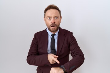 Middle age business man with beard wearing suit and tie in hurry pointing to watch time,...