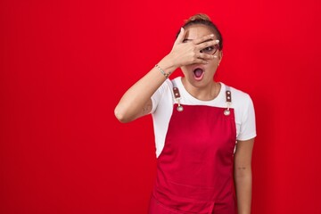 Young hispanic woman wearing waitress apron over red background peeking in shock covering face and eyes with hand, looking through fingers with embarrassed expression.