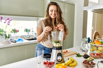 Young beautiful hispanic woman preparing vegetable smoothie with blender at the kitchen