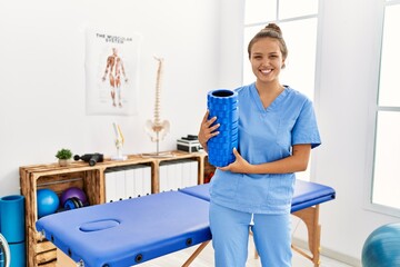 Young beautiful hispanic woman physiotherapist smiling confident holding foam roller at rehab clinic