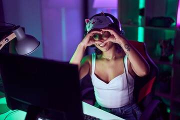 Young beautiful hispanic woman streamer smiling confident doing heart symbol with hands at gaming room