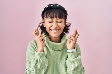 Young beautiful woman standing over pink background gesturing finger crossed smiling with hope and eyes closed. luck and superstitious concept.