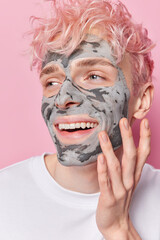 Vertical shot of cheerful teenage man applies facial clay mask to reduce pores and acnes touches face gently smiles brroadly looks happily aside wears white t shirt isolated over pink background.