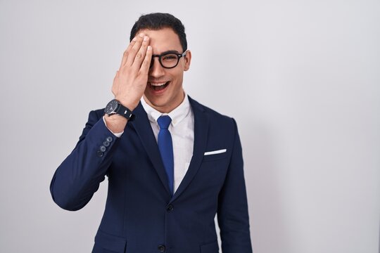 Young hispanic man wearing suit and tie covering one eye with hand, confident smile on face and surprise emotion.
