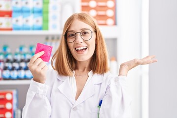 Young redhead woman working at pharmacy drugstore holding condom celebrating victory with happy smile and winner expression with raised hands