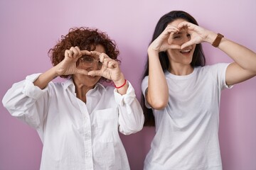 Hispanic mother and daughter wearing casual white t shirt over pink background doing heart shape with hand and fingers smiling looking through sign