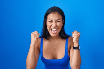 Hispanic woman standing over blue background angry and mad raising fists frustrated and furious while shouting with anger. rage and aggressive concept.