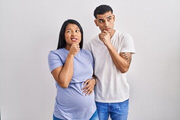 Young hispanic couple expecting a baby standing over background thinking worried about a question, concerned and nervous with hand on chin