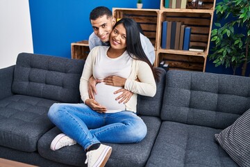 Obraz premium Young latin couple expecting baby hugging each other sitting on sofa at home