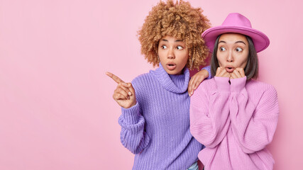 Horizontal shot of impressed two fashionable women stand speechless point index finger aside on blank space wear knitted jumpers hat isolated on pink background copy space for your promotional content