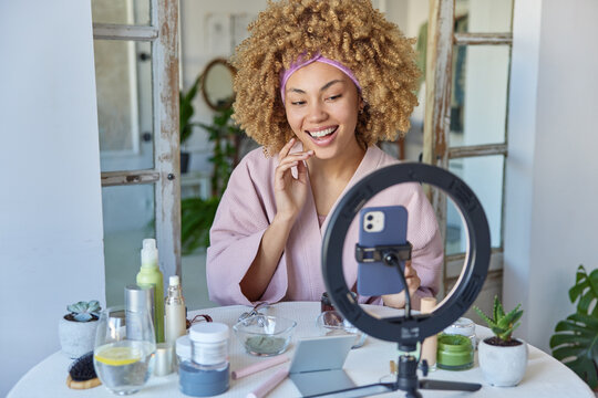 Beautiful smiling young woman with curly hair dressed in robe records video for her beauty blog sits at table with various cosmetic products has healthy skin poses against domestic interior.