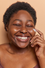 Headshot of cheerful dark skinned woman applies cosmetic cream on face keeps eyes closed enjoys...