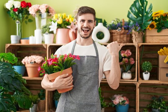 Hispanic man with beard working at florist shop holding plant pointing thumb up to the side smiling happy with open mouth