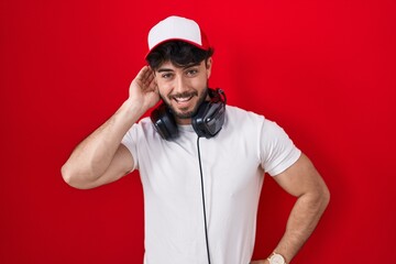 Hispanic man with beard wearing gamer hat and headphones smiling with hand over ear listening an hearing to rumor or gossip. deafness concept.