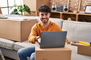 Young hispanic man smiling confident using laptop at new home - obrazy, fototapety, plakaty