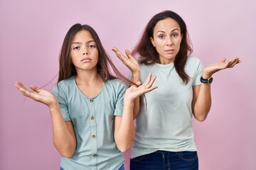 Young mother and daughter standing over pink background clueless and confused expression with arms and hands raised. doubt concept.