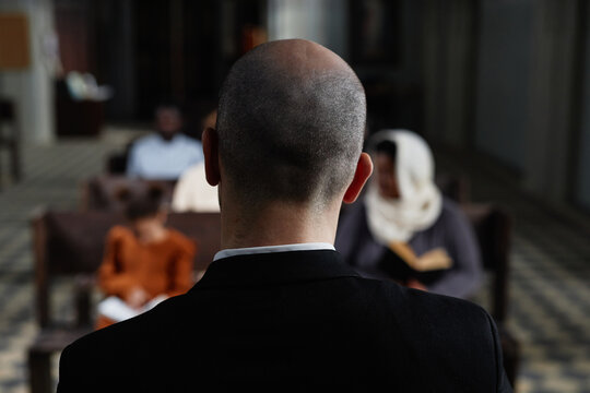 Rear view of protestant in black suit reading Bible for believers while they sitting on bench in baptist church