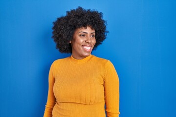 Fototapeta na wymiar Black woman with curly hair standing over blue background looking away to side with smile on face, natural expression. laughing confident.