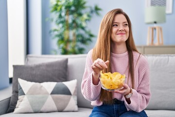 Young caucasian woman eating chips potatoes sitting on sofa at home - Powered by Adobe