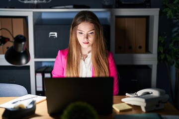 Young caucasian woman working at the office at night relaxed with serious expression on face. simple and natural looking at the camera.
