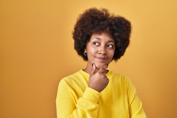 Fototapeta na wymiar Young african american woman standing over yellow background looking confident at the camera smiling with crossed arms and hand raised on chin. thinking positive.