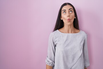 Young brunette woman standing over pink background making fish face with lips, crazy and comical gesture. funny expression.