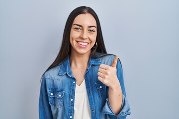 Hispanic woman standing over blue background doing happy thumbs up gesture with hand. approving...
