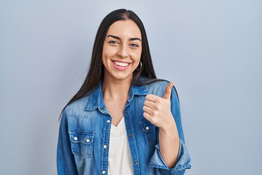 Hispanic woman standing over blue background doing happy thumbs up gesture with hand. approving expression looking at the camera showing success.