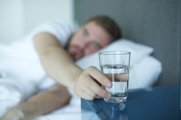 Young handsome man in the bed in bedroom at home in the morning lying under white blanket and drinking pure fresh water from glass 