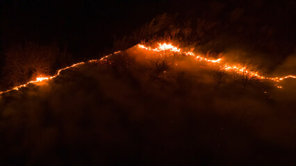 Night fire in the forest with fire and smoke.Epic aerial photo of a smoking wild flame.A blazing,glowing fire at night.Forest fires.Dry grass is burning. climate change,ecology.Line fire in the dark.