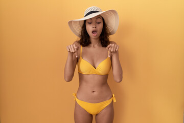 Young hispanic woman wearing bikini and summer hat pointing down with fingers showing advertisement, surprised face and open mouth