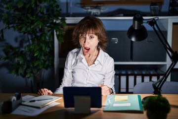 Middle age woman working at the office at night in shock face, looking skeptical and sarcastic, surprised with open mouth