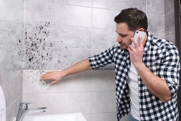 Mold removal service. Man talking on phone and looking at affected walls in bathroom