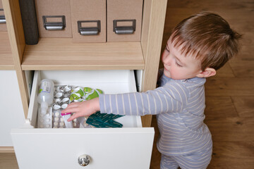 Toddler baby opened the cabinet drawer with pills and medicine. Child boy holding a pack of pills in the home living room. Kid age one year nine months - obrazy, fototapety, plakaty