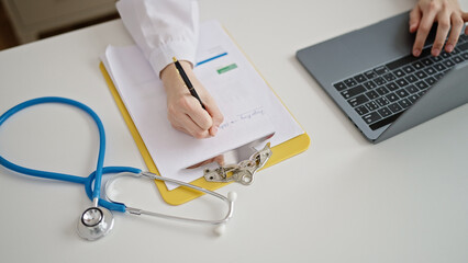 Young beautiful hispanic woman doctor using laptop writing on clipboard at clinic