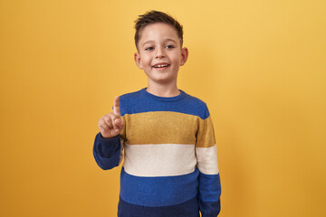 Little hispanic boy standing over yellow background smiling with an idea or question pointing finger up with happy face, number one