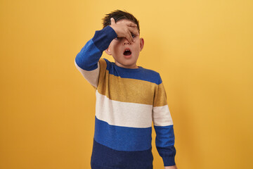 Little hispanic boy standing over yellow background peeking in shock covering face and eyes with...