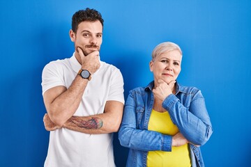Young brazilian mother and son standing over blue background looking confident at the camera smiling with crossed arms and hand raised on chin. thinking positive.