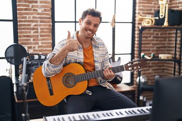 Young hispanic man playing classic guitar at music studio smiling happy and positive, thumb up...