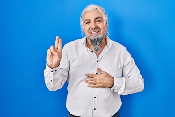 Middle age man with grey hair standing over blue background smiling swearing with hand on chest and...